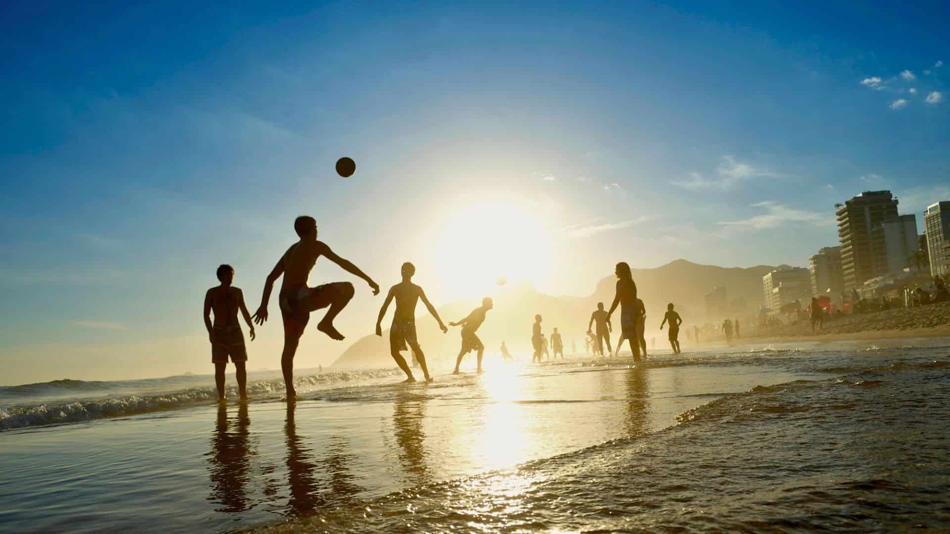 People play soccer on a beach at sunset, with buildings and mountains in the background.