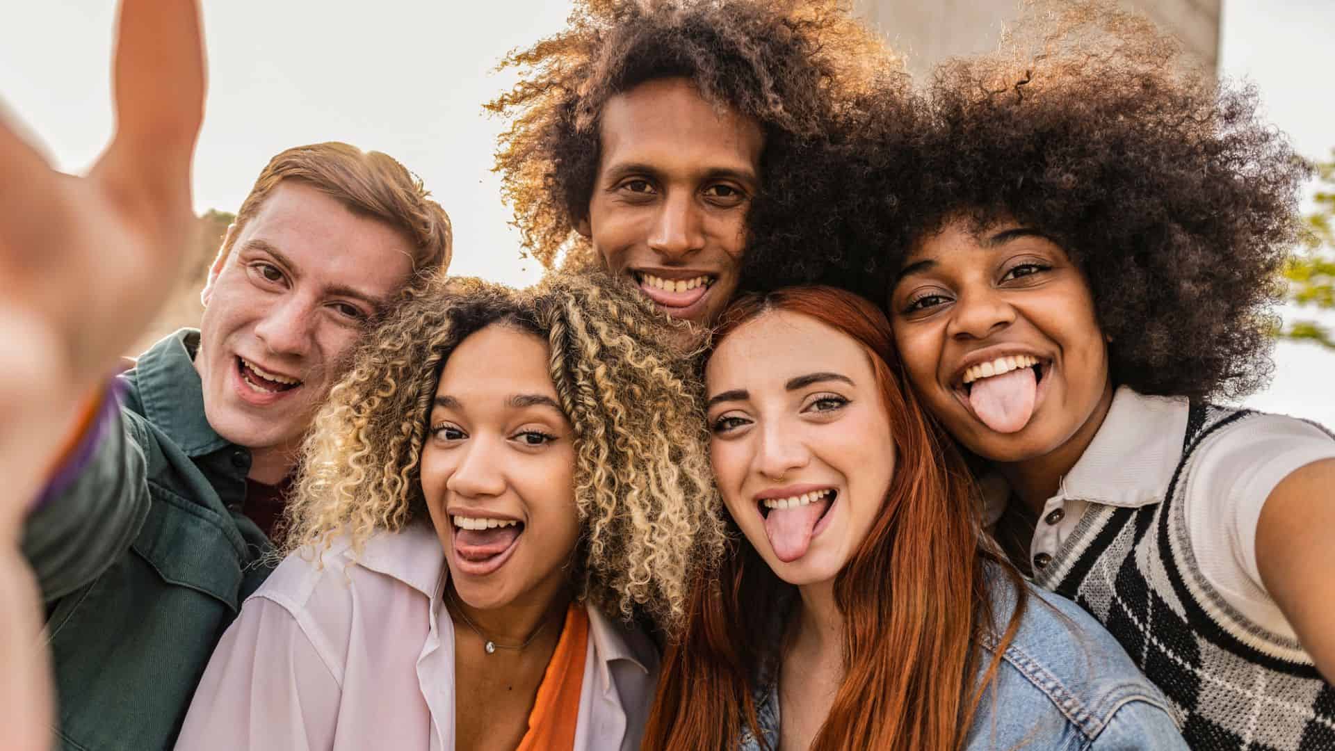 Five friends smile and make silly faces while taking a selfie outdoors on a sunny day.