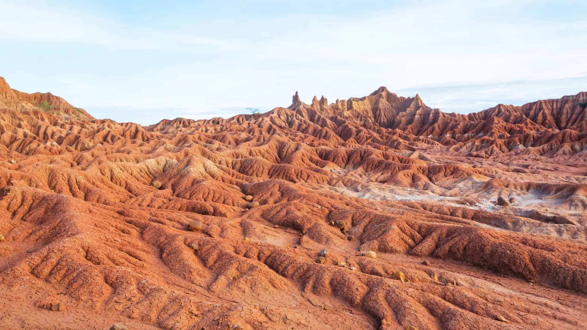 Red, rocky desert landscape with ridges and peaks under a bright, partly cloudy sky.