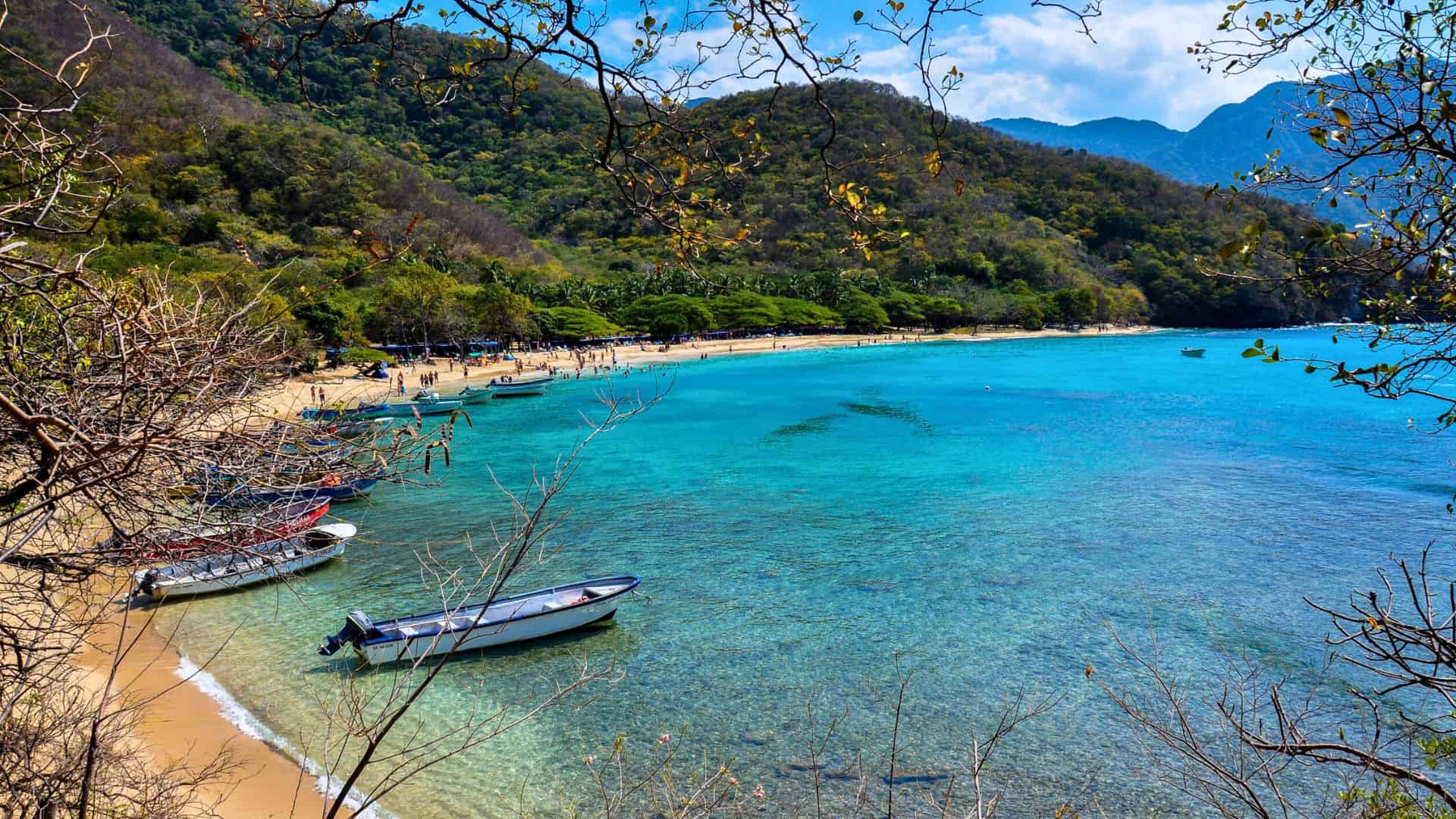 Clear turquoise water, boats near a sandy beach, and lush green hills under a partly cloudy sky.