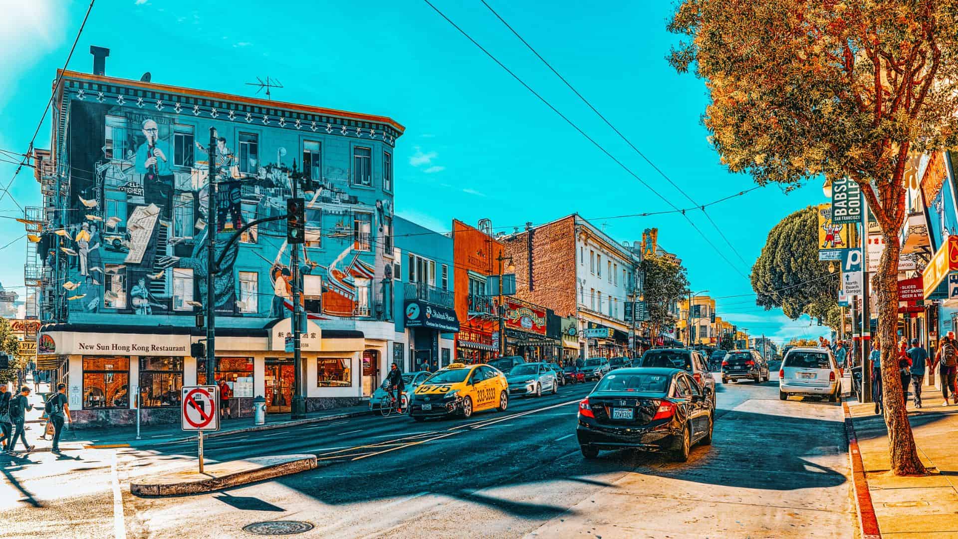 Colorful city street with cars, pedestrians, and a large mural on a building under a bright blue sky.