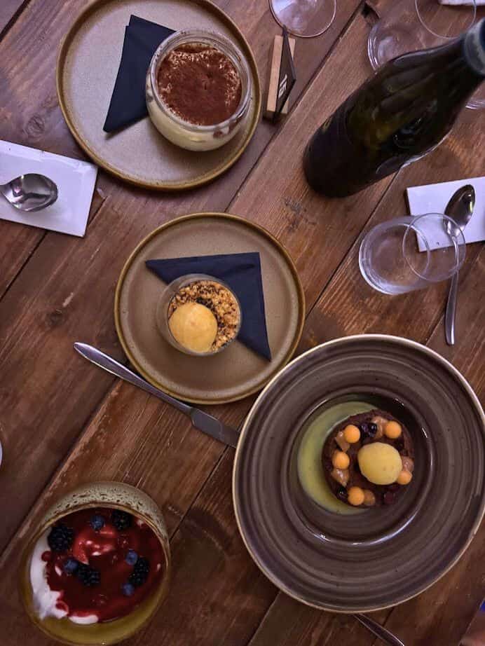 Four assorted plated desserts on a rustic wooden table evoke the charm of 4 days in Castel Gandolfo, complete with glasses, utensils, and a wine bottle for a perfect Italian escape.
