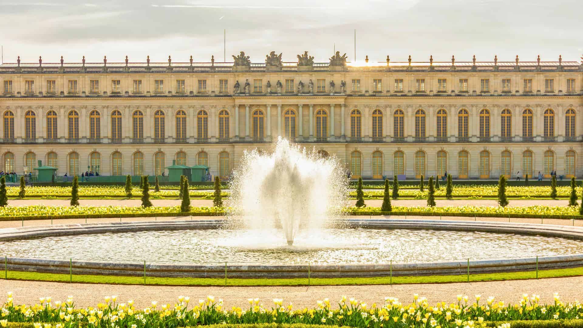 A large fountain in front of the Palace of Versailles, surrounded by gardens and blooming flowers.