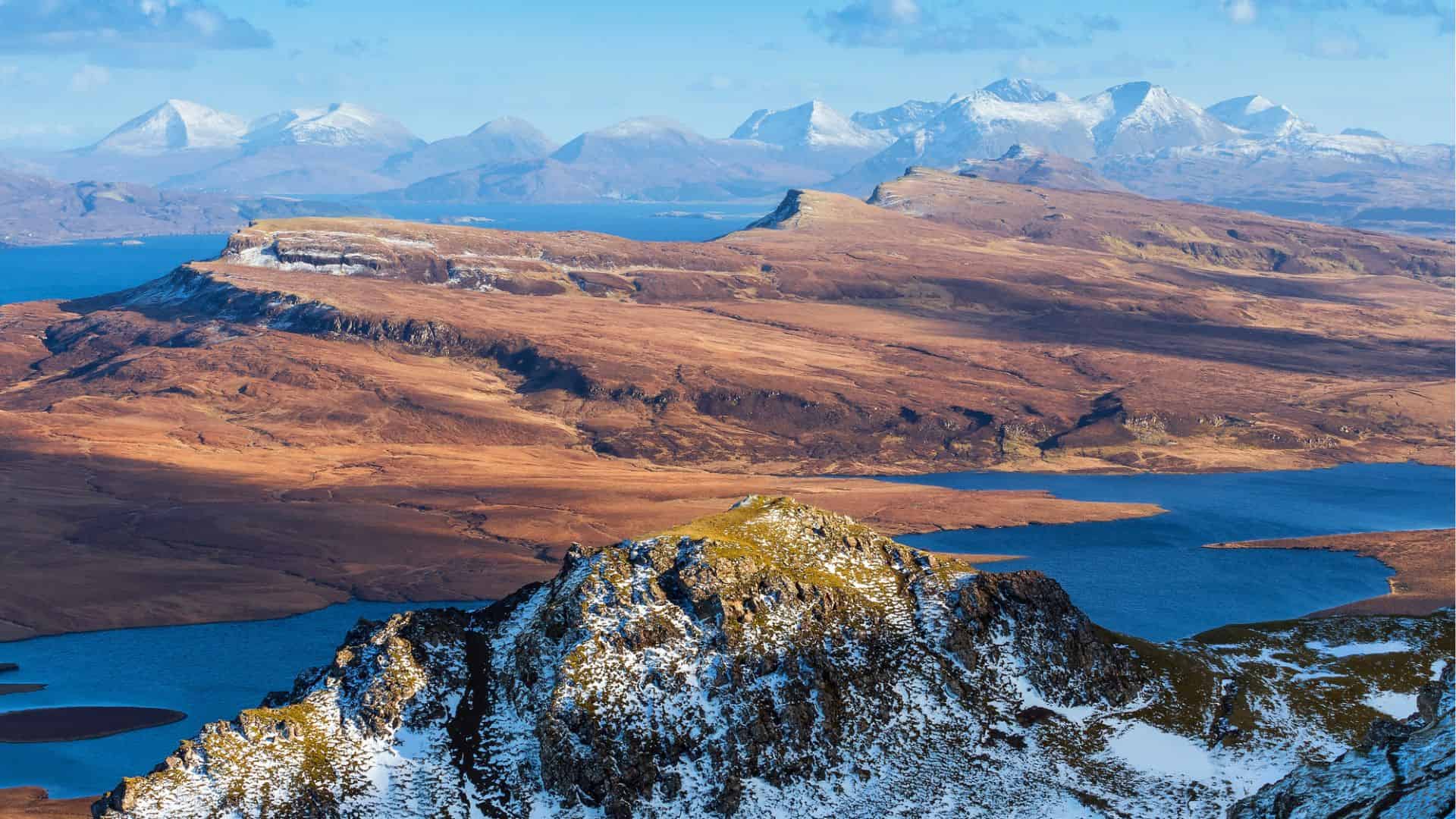 Snow-capped mountains, brown hills, and blue lakes under a clear sky in a scenic landscape.