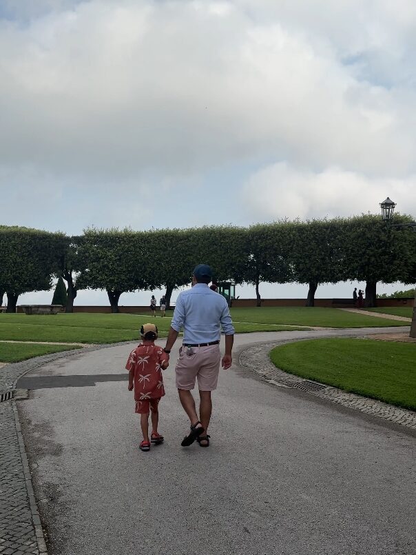 An adult and child walk hand in hand on a tree-lined path in a park under a cloudy sky, reminiscing about their 4 days in Castel Gandolfo.