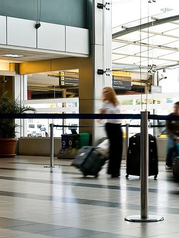 People with suitcases walking through an airport terminal near the entrance, cars visible outside through glass doors.