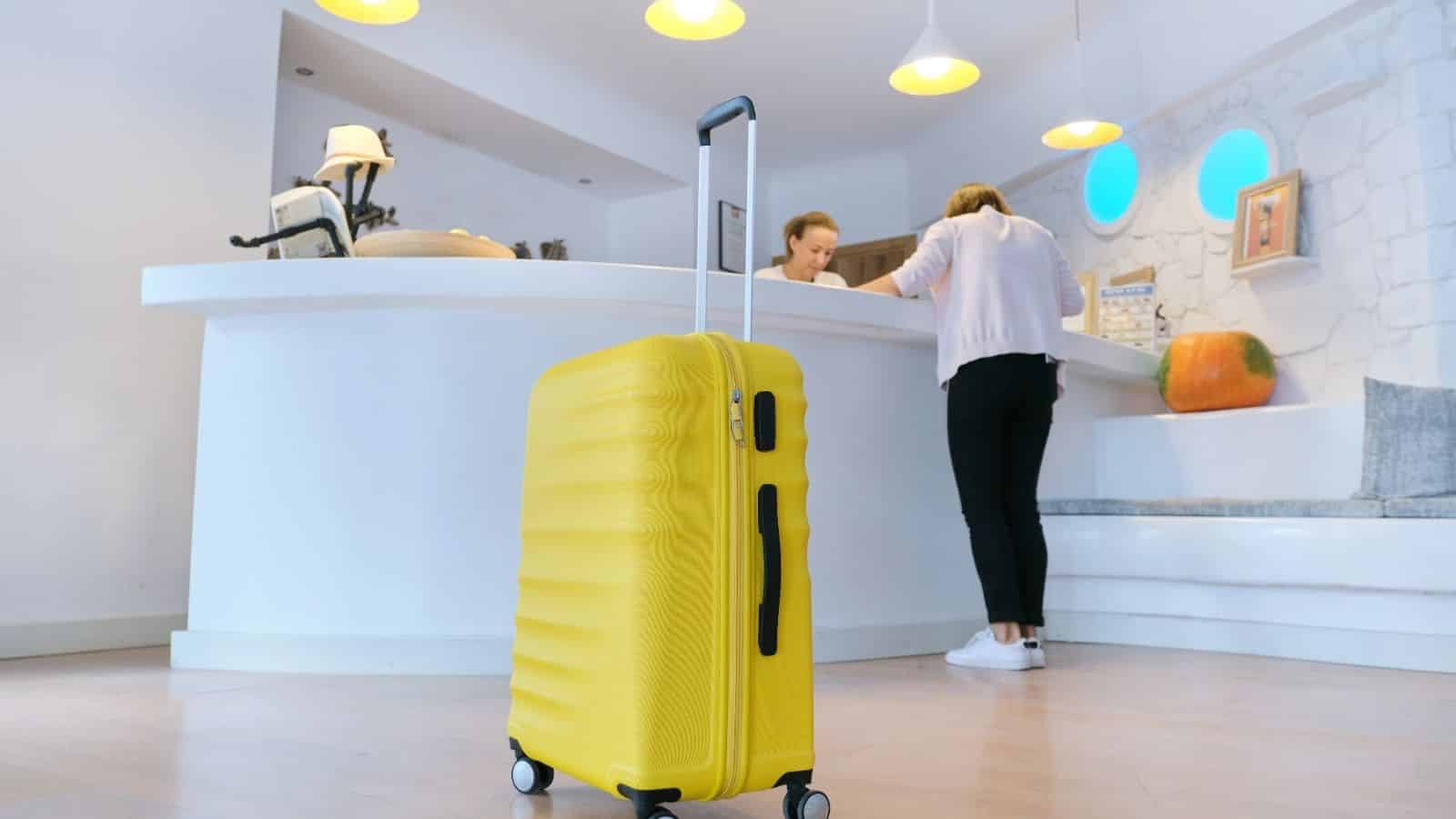 A yellow suitcase stands in a hotel lobby near a reception desk where two people are checking in.