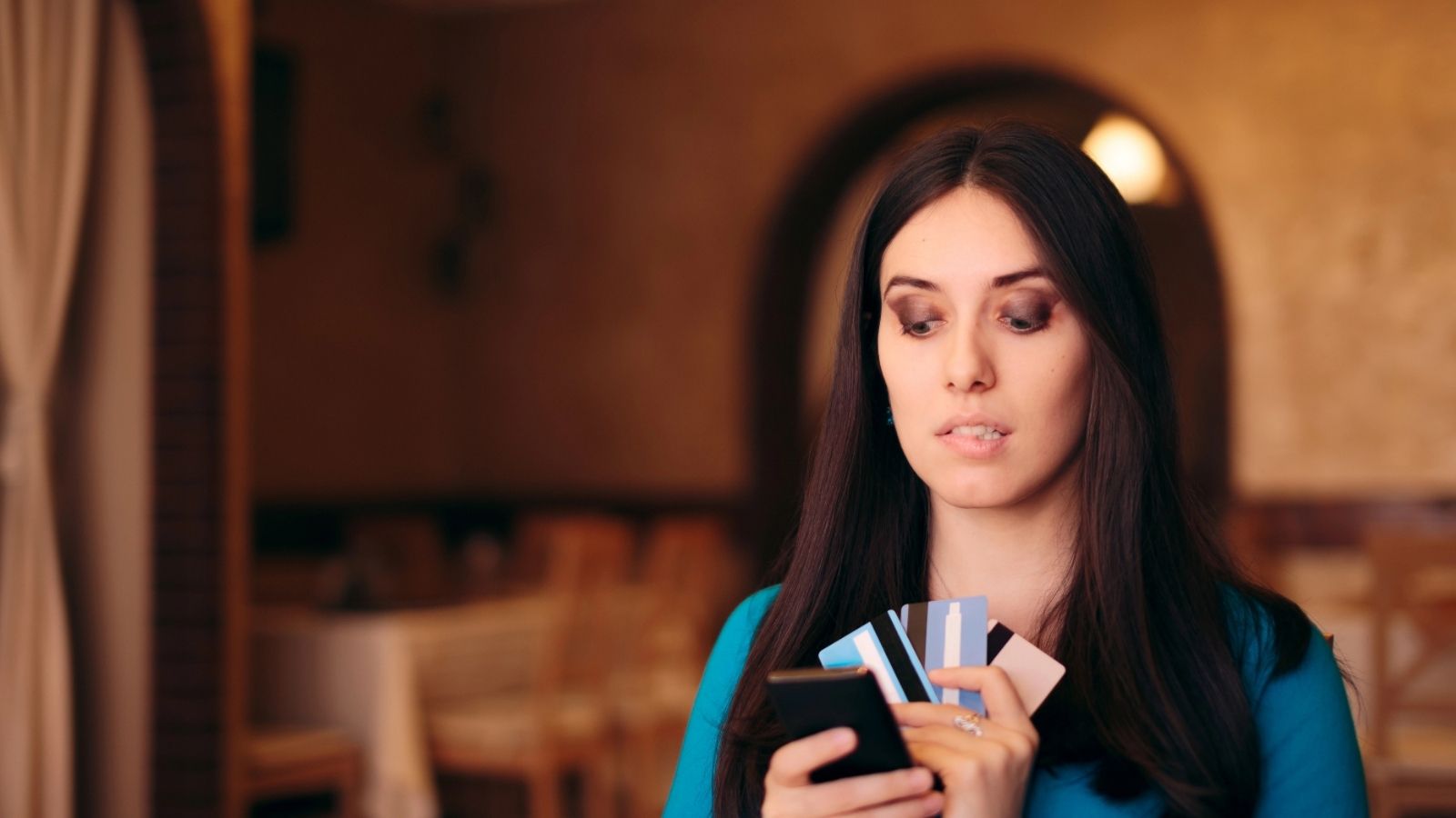 Woman holding several credit cards and looking at her phone in a softly lit indoor setting.
