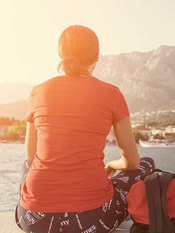 A person sits by the water with a backpack, looking at a coastal town and mountains in the distance.
