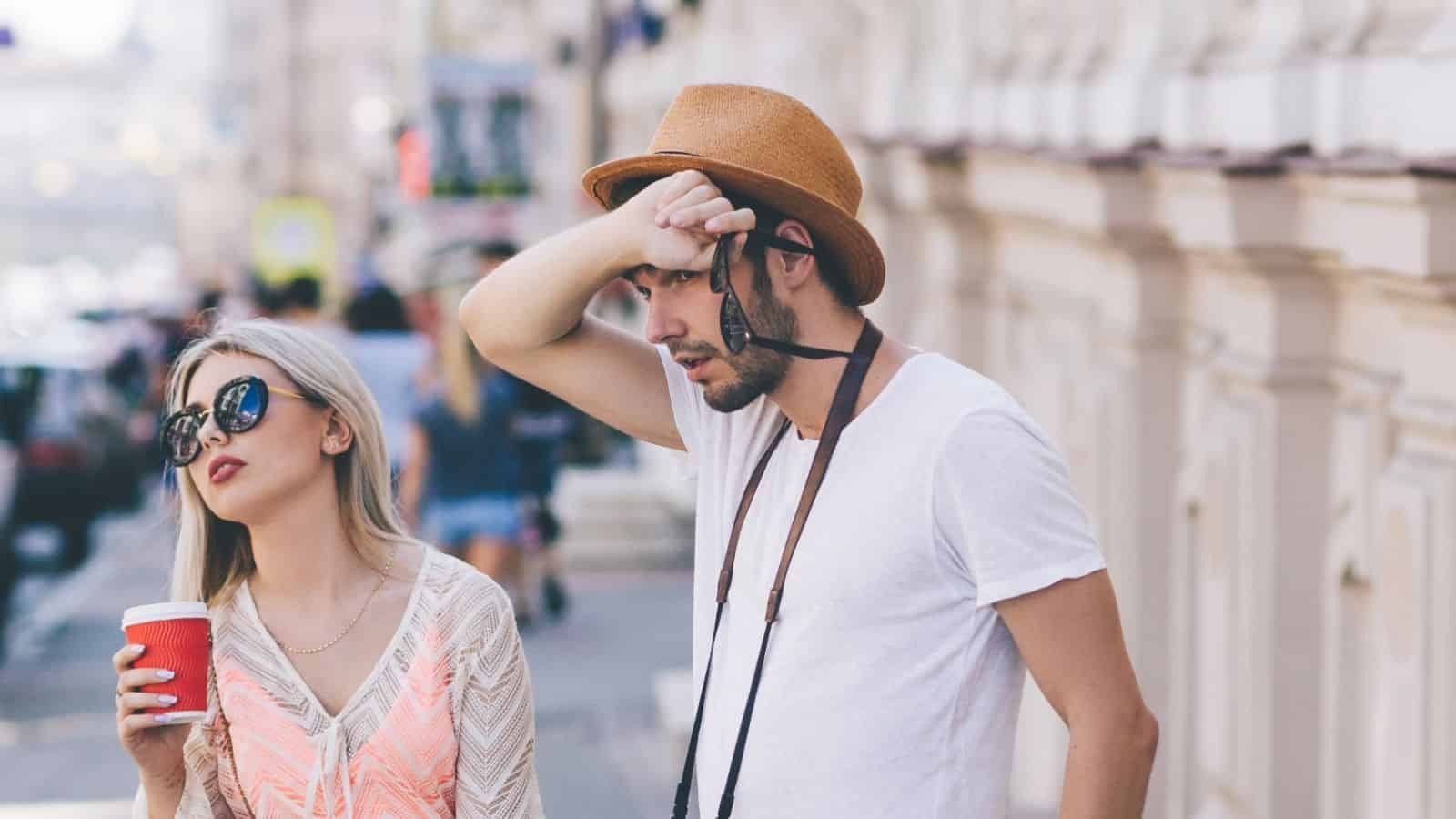 Man in hat wipes forehead while woman with sunglasses holds coffee cup, both walking down a sunny city street.
