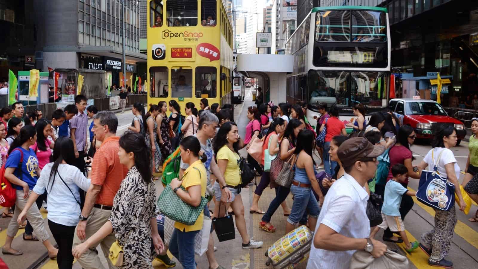 Crowd of people crossing a busy street in a city, with trams, cars, and tall buildings in the background.