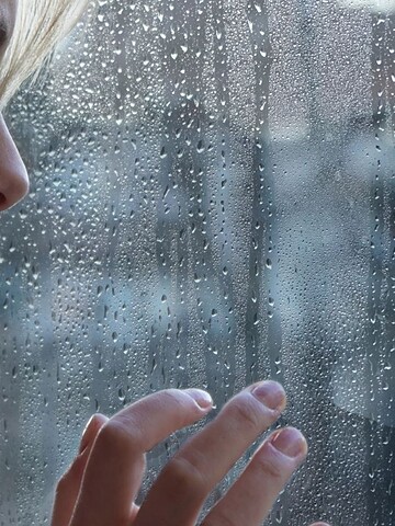 A woman looks thoughtfully out a rain-covered window, touching the glass with her fingers.