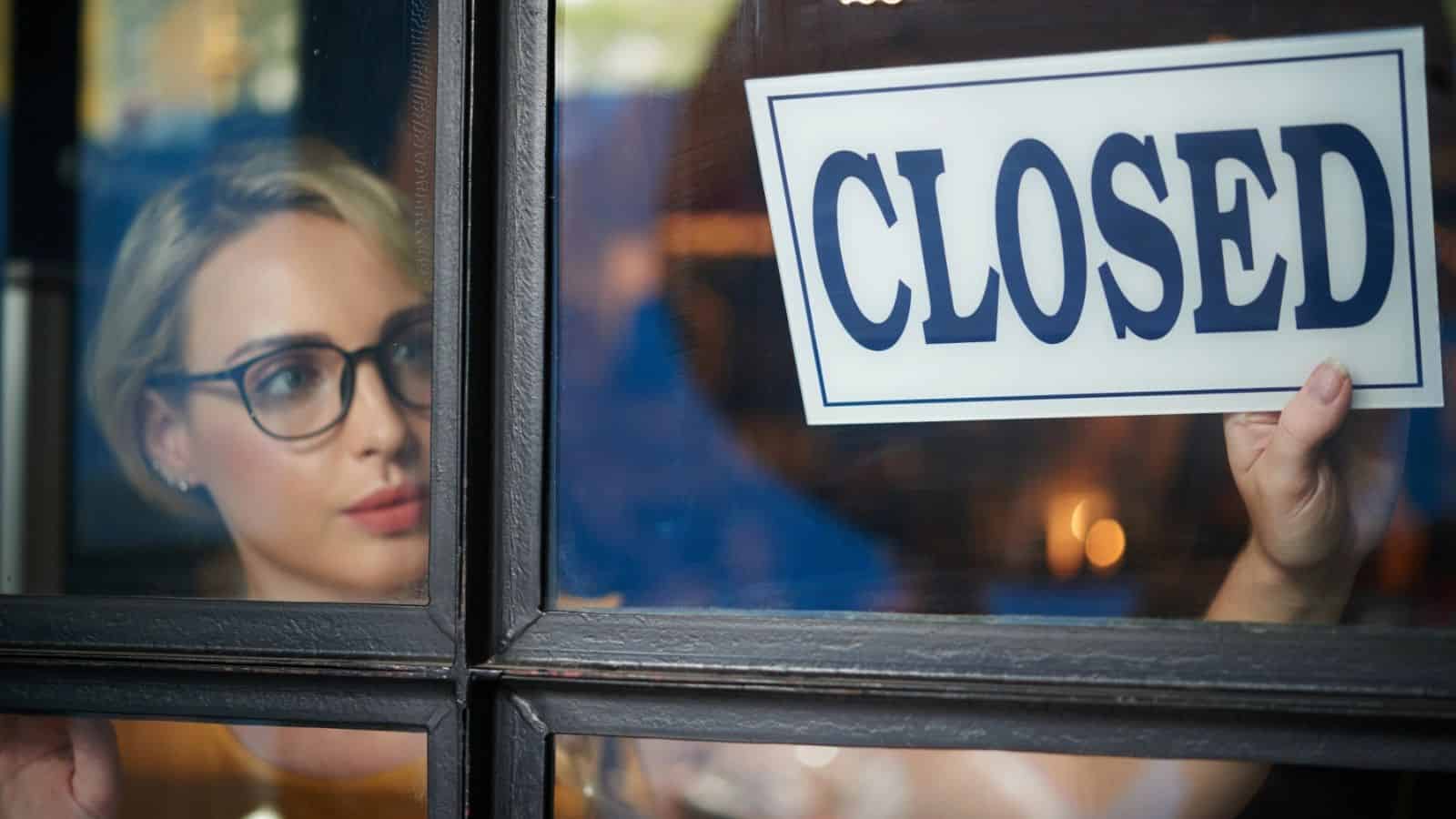A person hangs a "CLOSED" sign on a glass door while looking outside through the window.