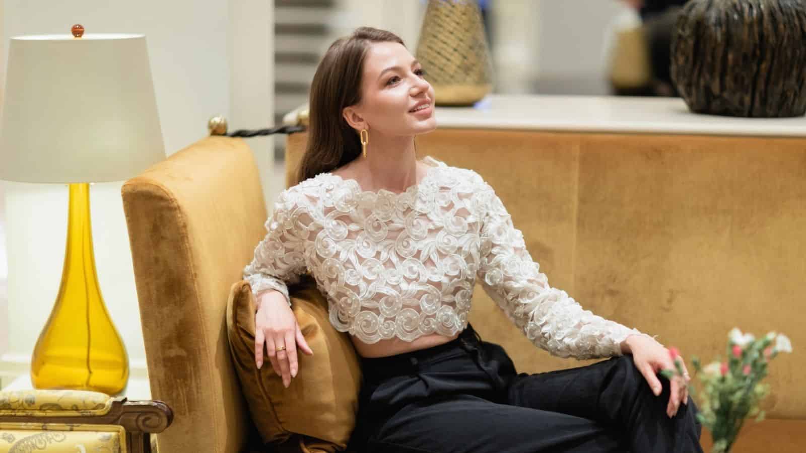 A woman in a white floral blouse and black pants sits on a yellow sofa, smiling and looking to the side.