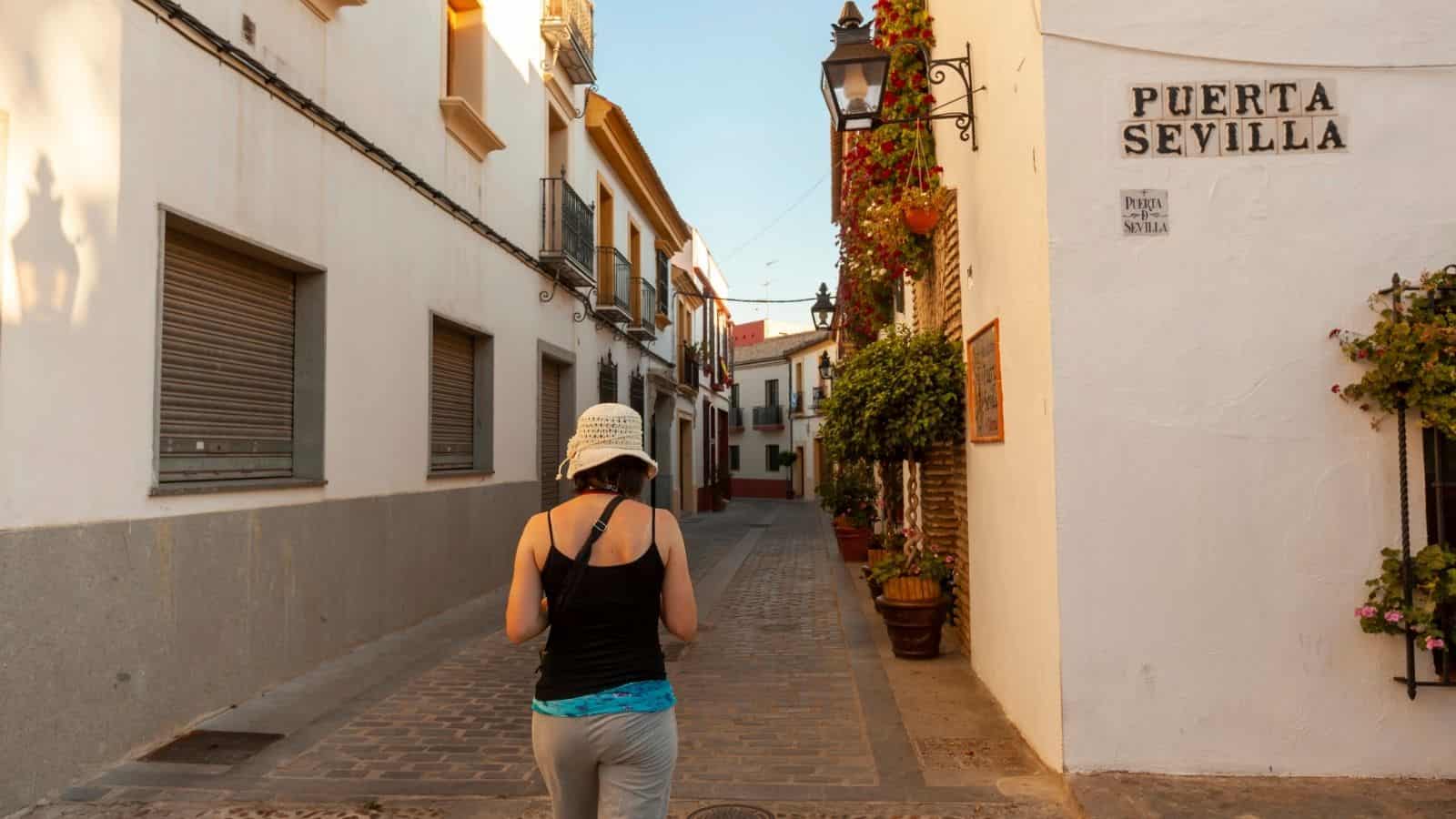 A woman in a white hat walks down a narrow, sunny street next to a building labeled "Puerta Sevilla.