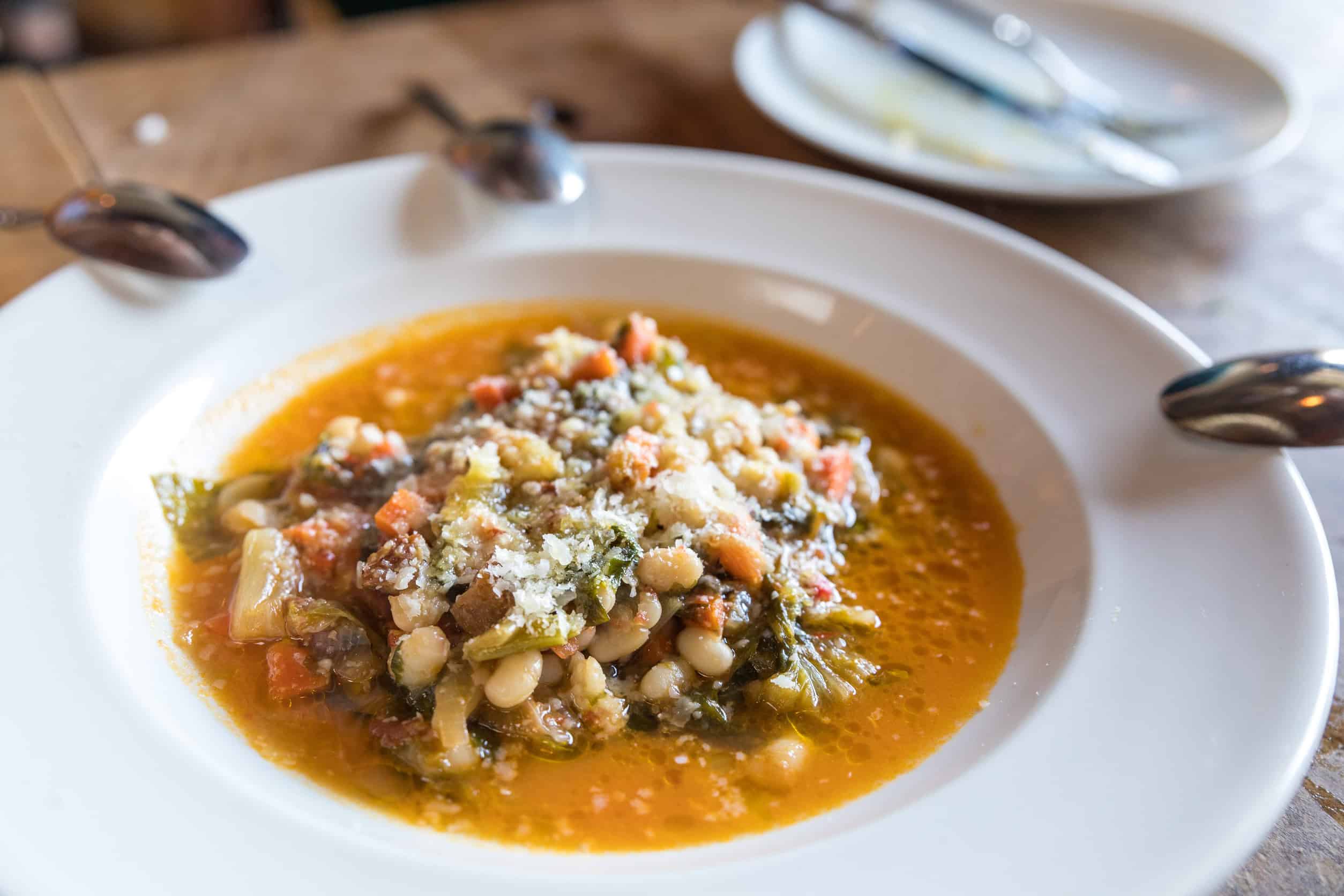 A bowl of vegetable and bean stew topped with grated cheese, served on a white plate&mdash;an inviting way to eat vegetarian in Italy.