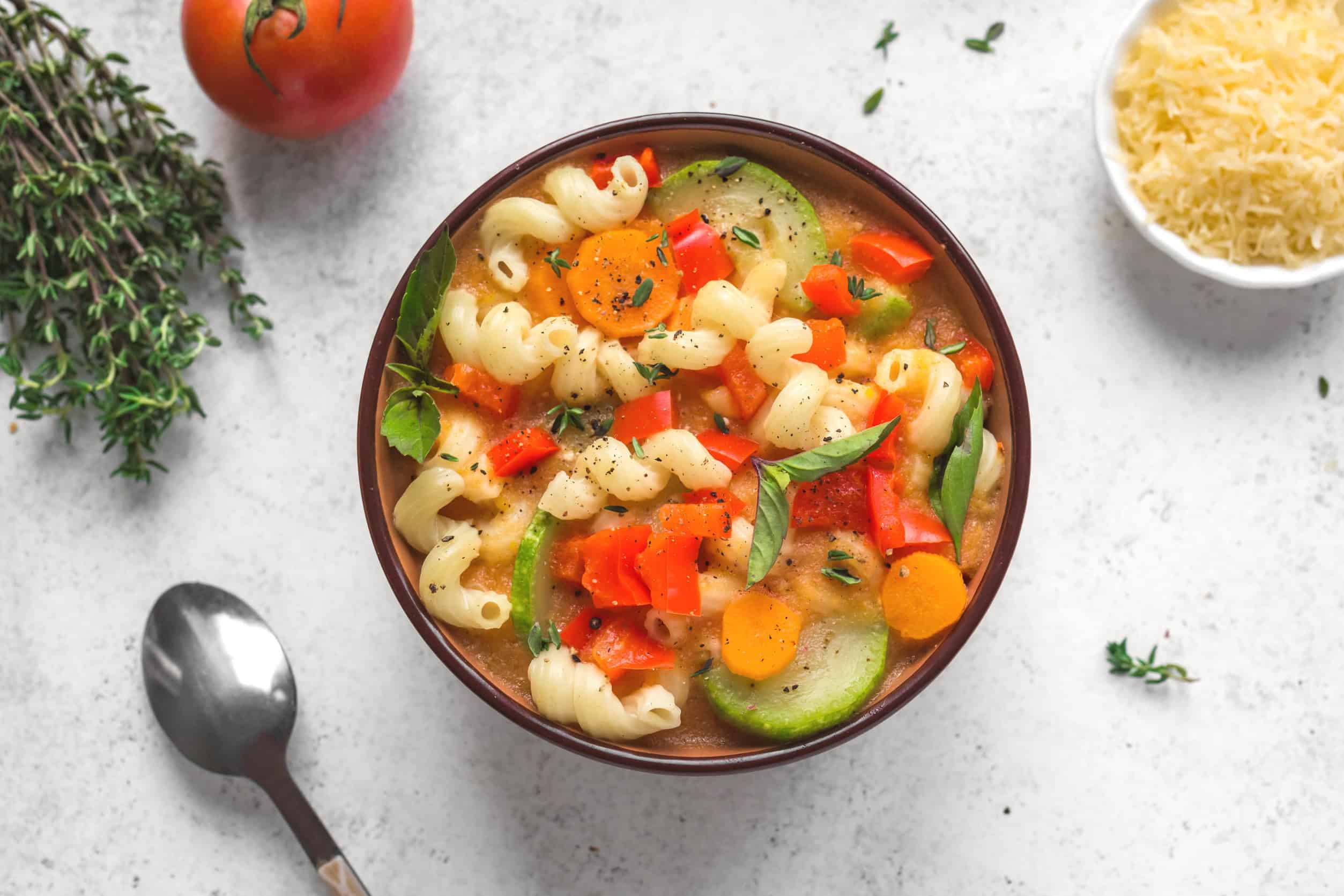 A bowl of vegetable soup with pasta, carrots, zucchini, and herbs&mdash;perfect for anyone looking to eat vegetarian in Italy&mdash;served alongside a spoon, cheese, and a fresh tomato.