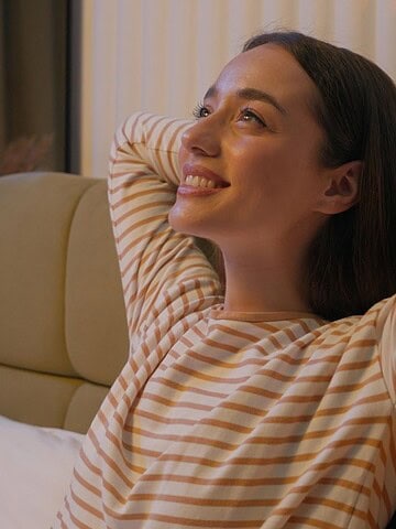 Woman in striped shirt smiles and relaxes with hands behind head while sitting on a bed in a cozy room.