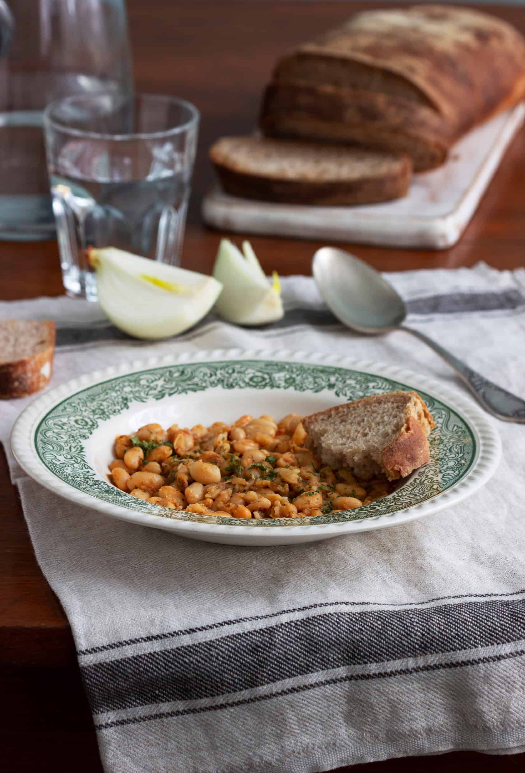 A plate of baked beans with bread, a glass of water, onion slices, and a loaf of bread in the background&mdash;an example of the Best Vegetarian Food in France.