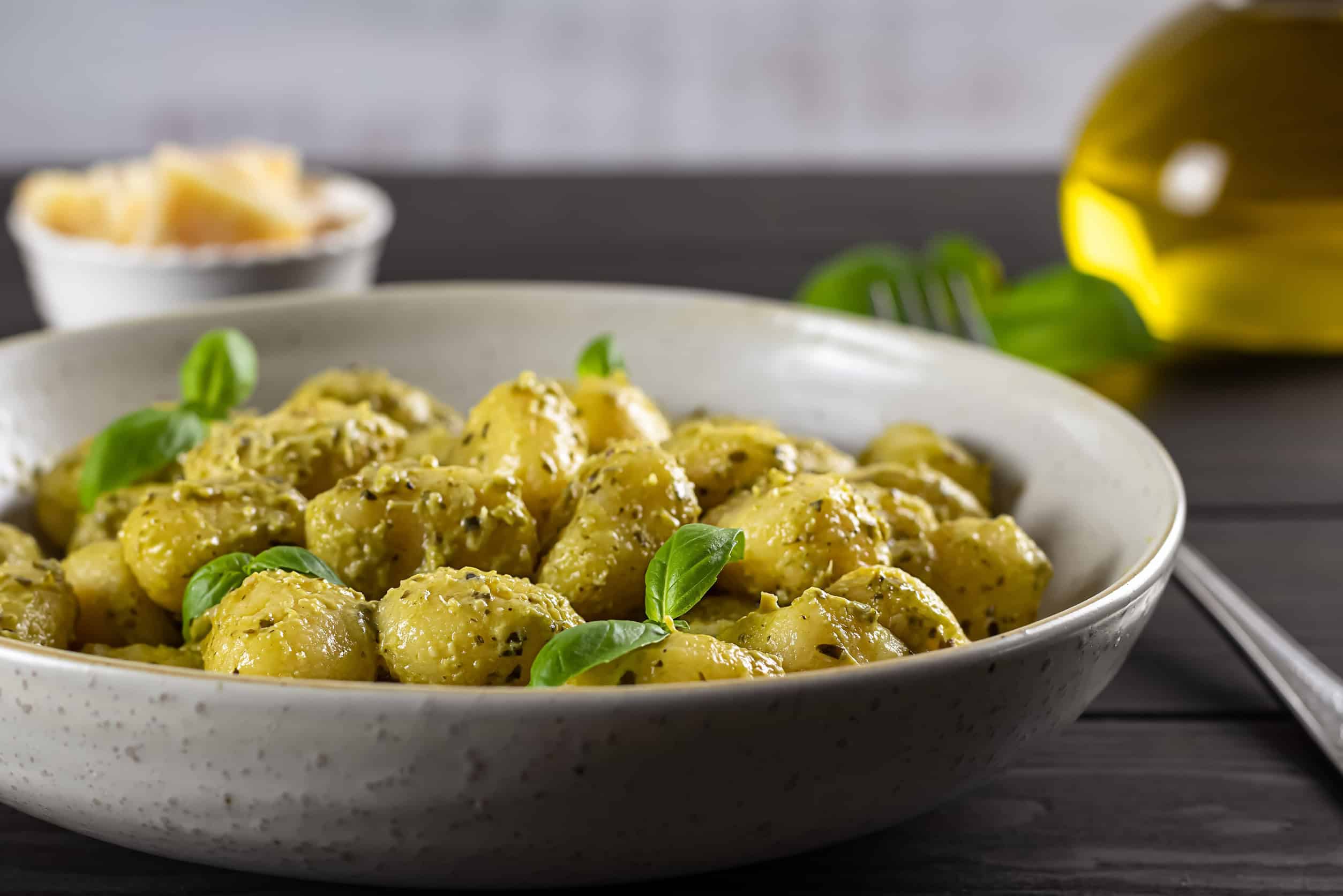 A bowl of gnocchi covered in pesto sauce, garnished with basil, showcases how easy it is to eat vegetarian in Italy, with cheese and olive oil in the background.