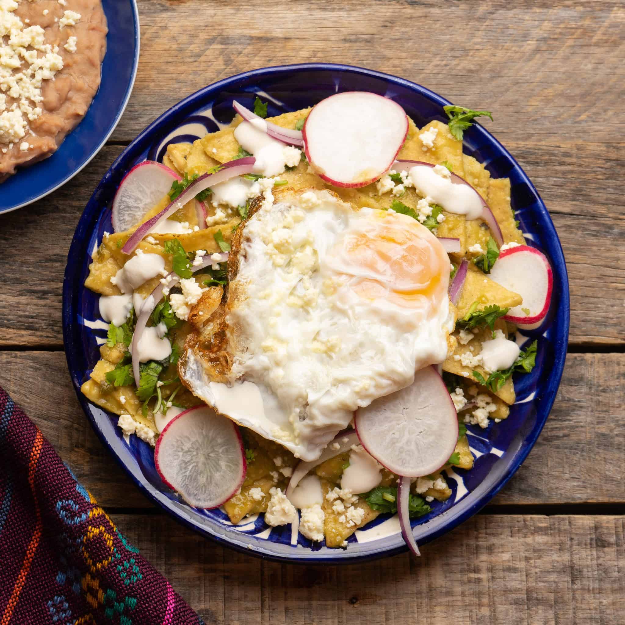 Plate of chilaquiles with fried egg, radish slices, cheese, crema, and herbs on a rustic wooden table&mdash;one of the best vegetarian Mexican dishes to enjoy any time of day.