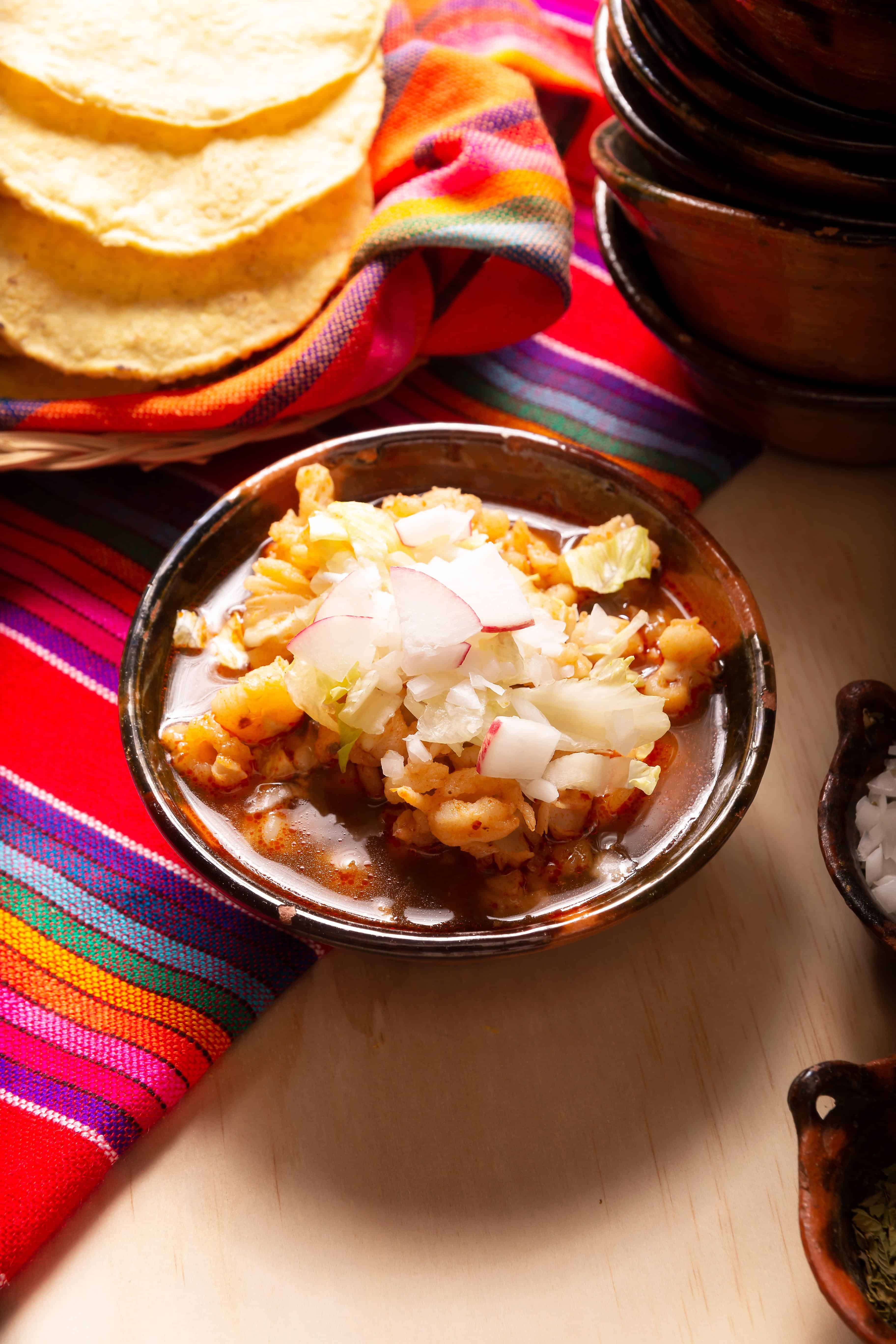A bowl of pozole, one of the best Vegetarian Mexican dishes, topped with chopped lettuce and radish sits beside tortillas on a colorful striped cloth.