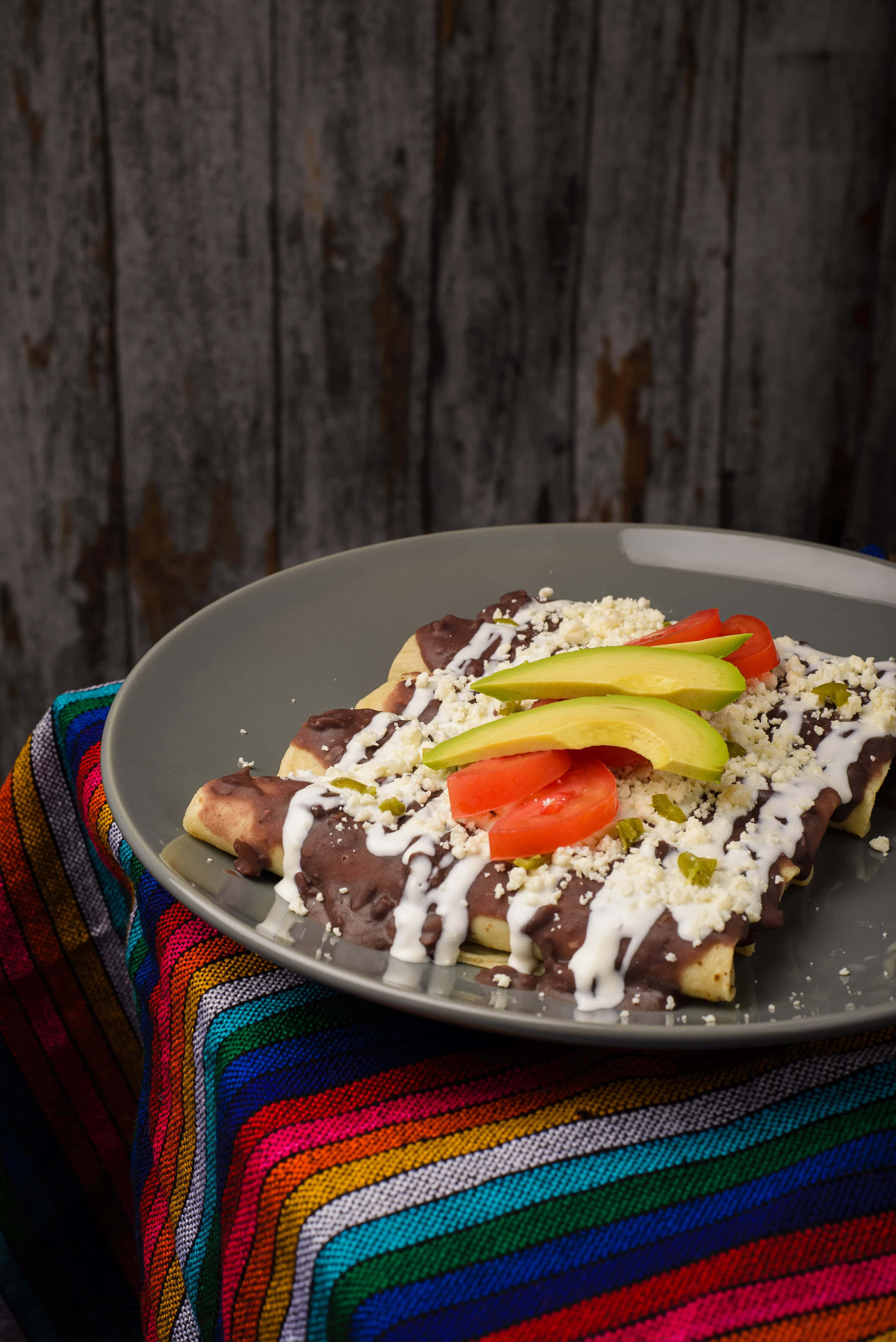 Enchiladas topped with black bean sauce, avocado, tomato, cheese, and crema on a gray plate with a colorful cloth&mdash;one of the best vegetarian Mexican dishes to savor.