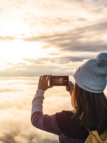 Person in a beanie takes a photo of a scenic sunrise above clouds and mountains with a smartphone.