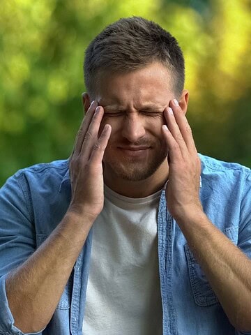 Man sitting on a bench outdoors, eyes closed, touching temples as if experiencing a headache or stress.