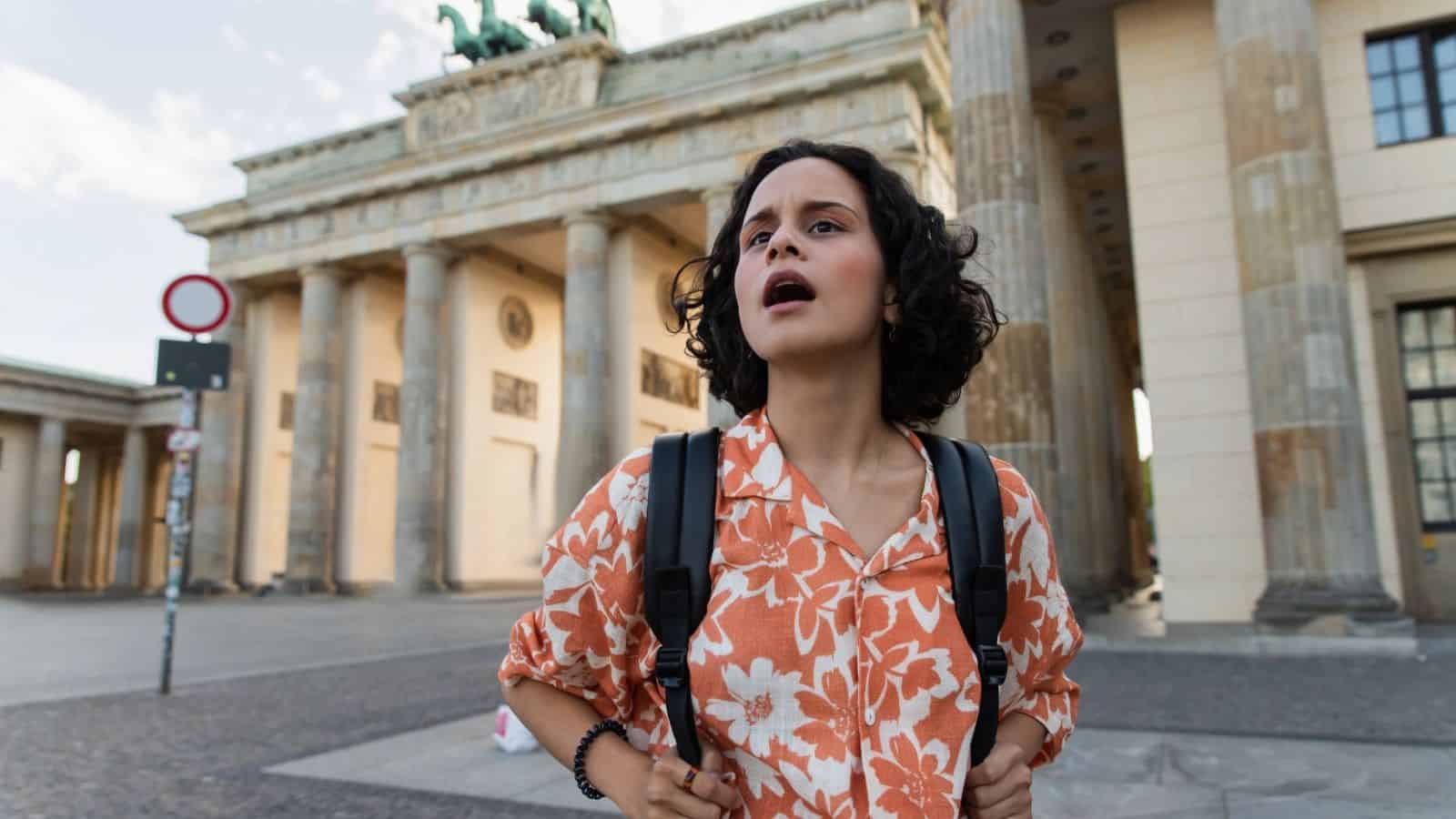 A woman with a backpack looks surprised in front of the Brandenburg Gate in Berlin, Germany.