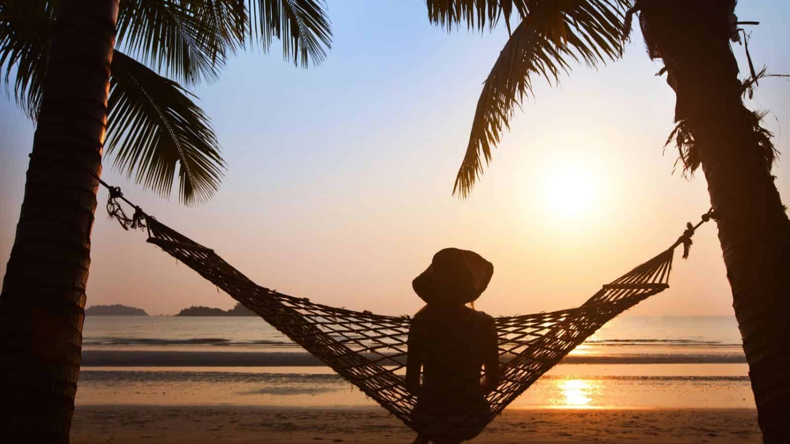 Person relaxing in a hammock between palm trees at the beach during sunset, facing the ocean.