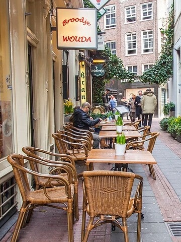 Narrow street with outdoor café seating, potted plants, and people walking in the background.