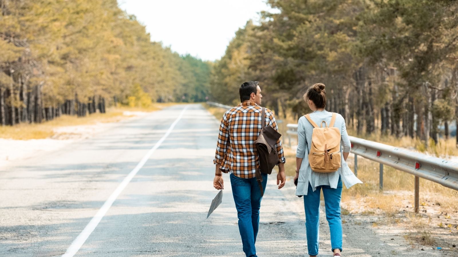 Two people with backpacks walk along a tree-lined road, talking and enjoying a sunny day outdoors.