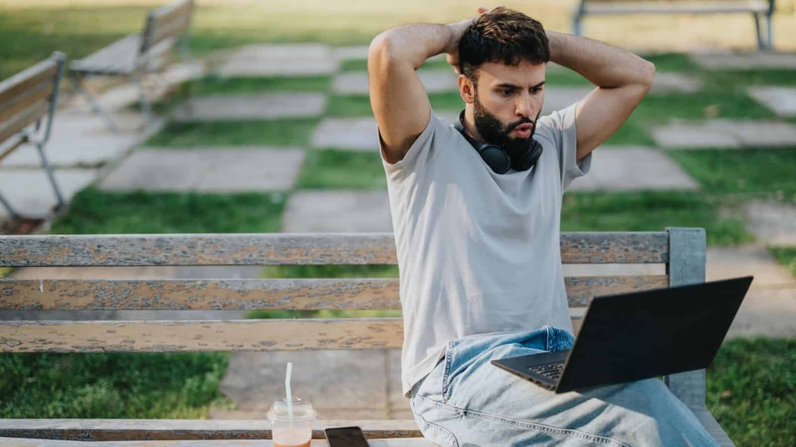 A surprised man with headphones looks at a laptop on a park bench, with a drink and phone beside him.