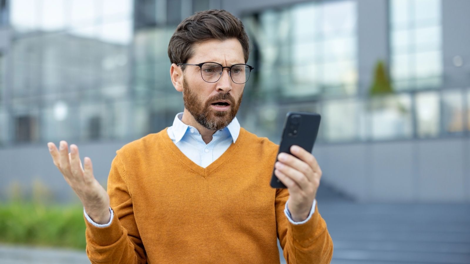 Man with glasses and orange sweater looks surprised at his smartphone while standing outdoors.