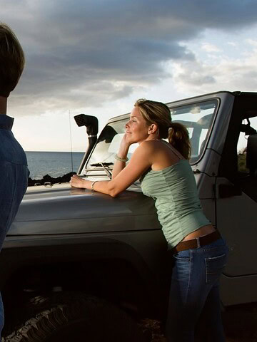 Man taking a photo by the ocean while a woman leans on a parked jeep, both enjoying the coastal view.