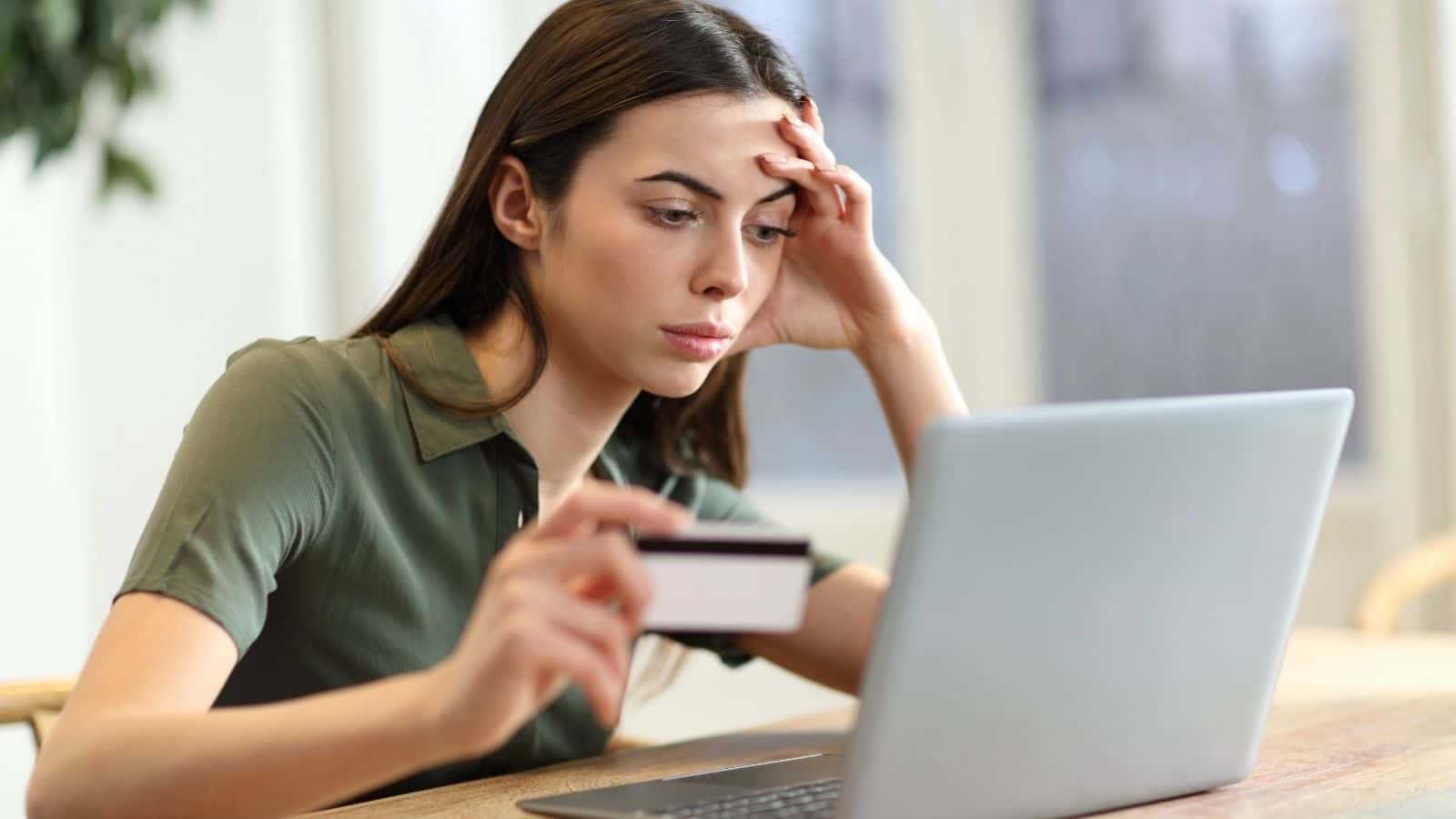 Woman holding a credit card and looking concerned while using a laptop at a desk.