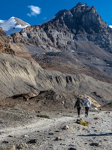 People hiking on a rocky trail with snow-capped mountains in the background under a clear blue sky.