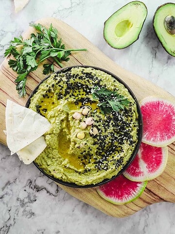 Bowl of green hummus topped with seeds, served with pita, parsley, radish slices, and avocado—a delicious example of Vegetarian Food in Israel.