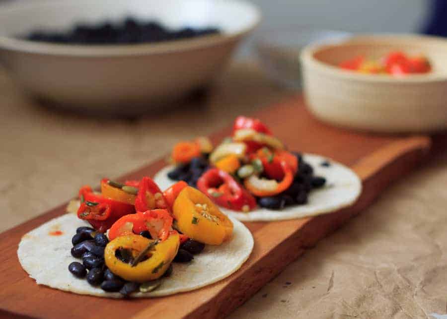 Two tortillas topped with black beans and sliced colorful peppers on a wooden board, these vibrant tacos are among the best vegetarian Mexican dishes, with bowls in the background adding extra flair.