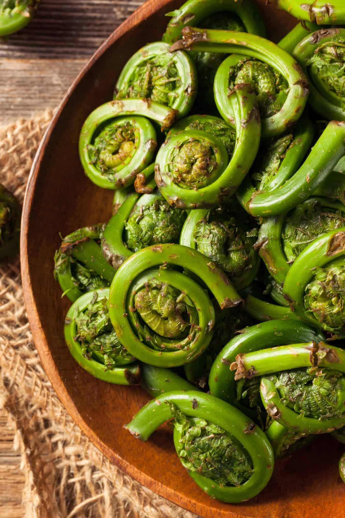 A wooden plate filled with cooked green fiddlehead ferns, a classic choice among Canadian Vegetarian Dishes, sits on a woven placemat.