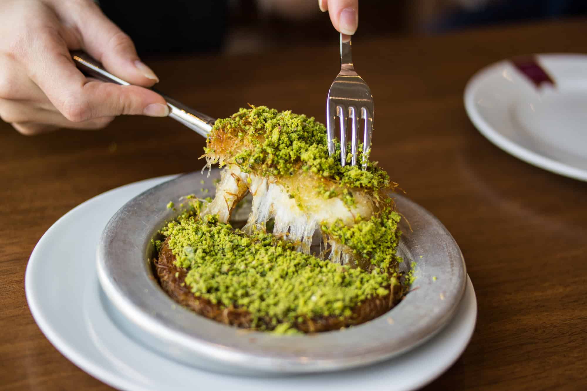 A person slices into a pistachio-covered kunafa dessert&mdash;an example of Vegetarian Food in Israel&mdash;on a metal plate with a fork and knife.