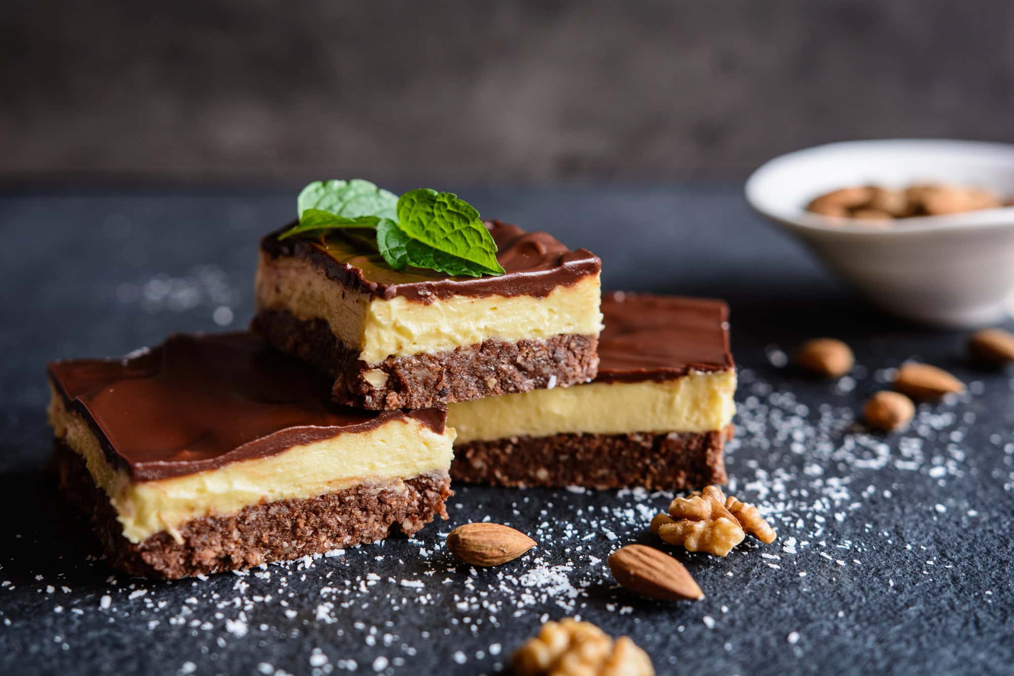 Three layered chocolate dessert bars, inspired by Canadian Vegetarian Dishes, topped with mint leaves, surrounded by nuts and a white bowl in the background.