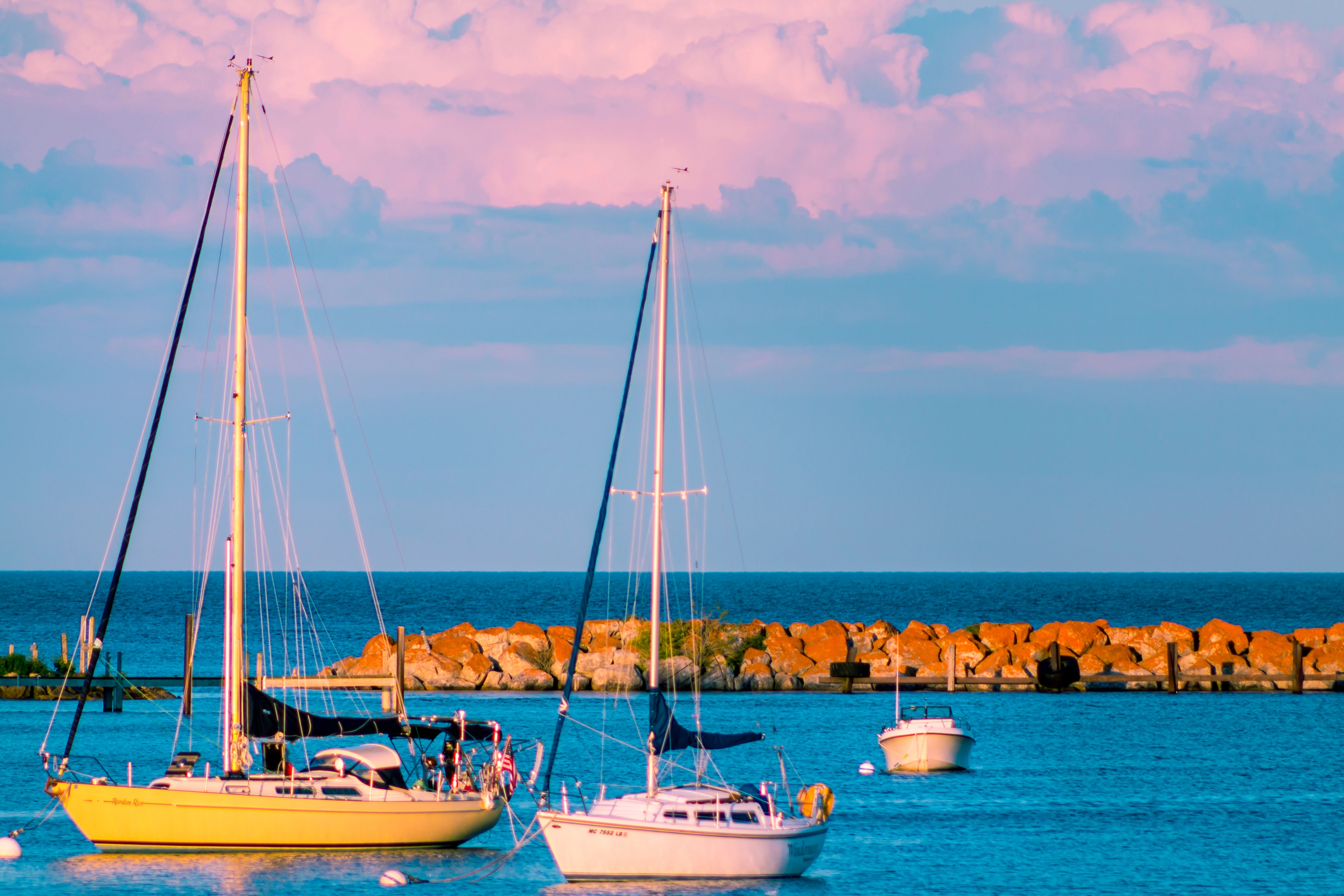Several sailboats and a motorboat float on calm blue water near a rocky breakwater under a pink-clouded sky&mdash;an idyllic scene perfect for family travel or anyone seeking bucket list travel destinations.