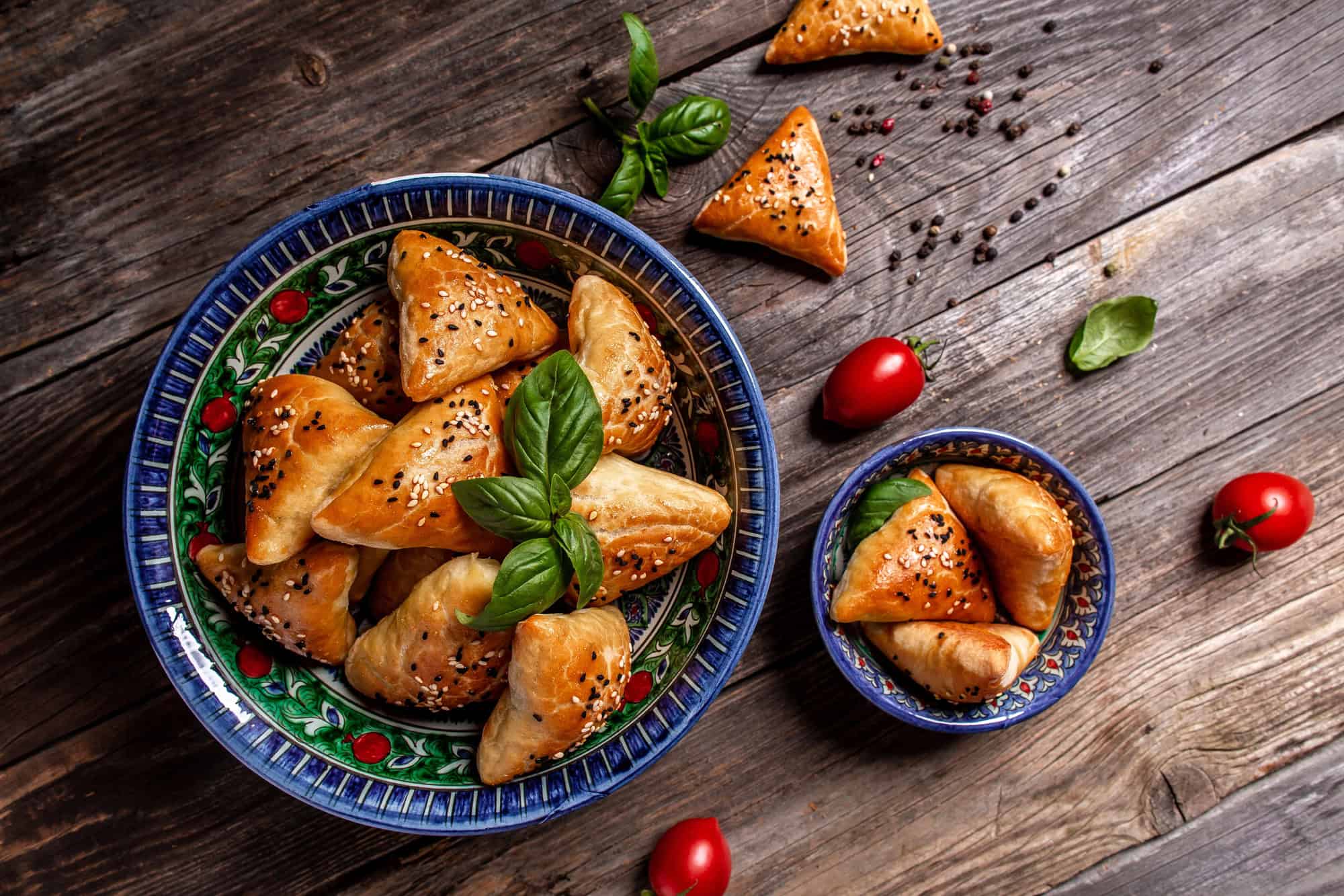 A bowl of golden, sesame-topped triangle pastries&mdash;a delicious example of Vegetarian Food in Israel&mdash;served with basil, tomatoes, and herbs on a rustic wooden table.