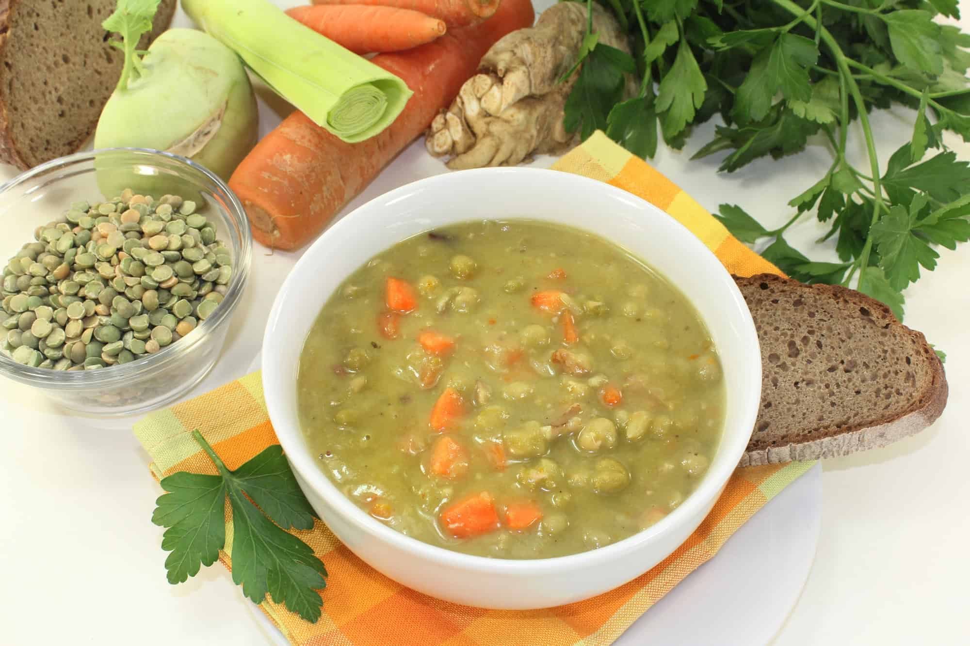 Bowl of pea soup with carrots, surrounded by bread, fresh vegetables, parsley, and a bowl of dried peas&mdash;a cozy favorite among Canadian Vegetarian Dishes.