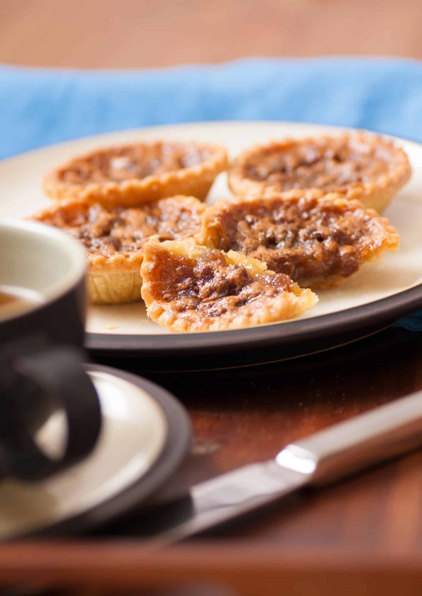 Five mini butter tarts, a staple among Canadian Vegetarian Dishes, rest on a plate beside a cup of tea and a knife, set atop a wooden table.