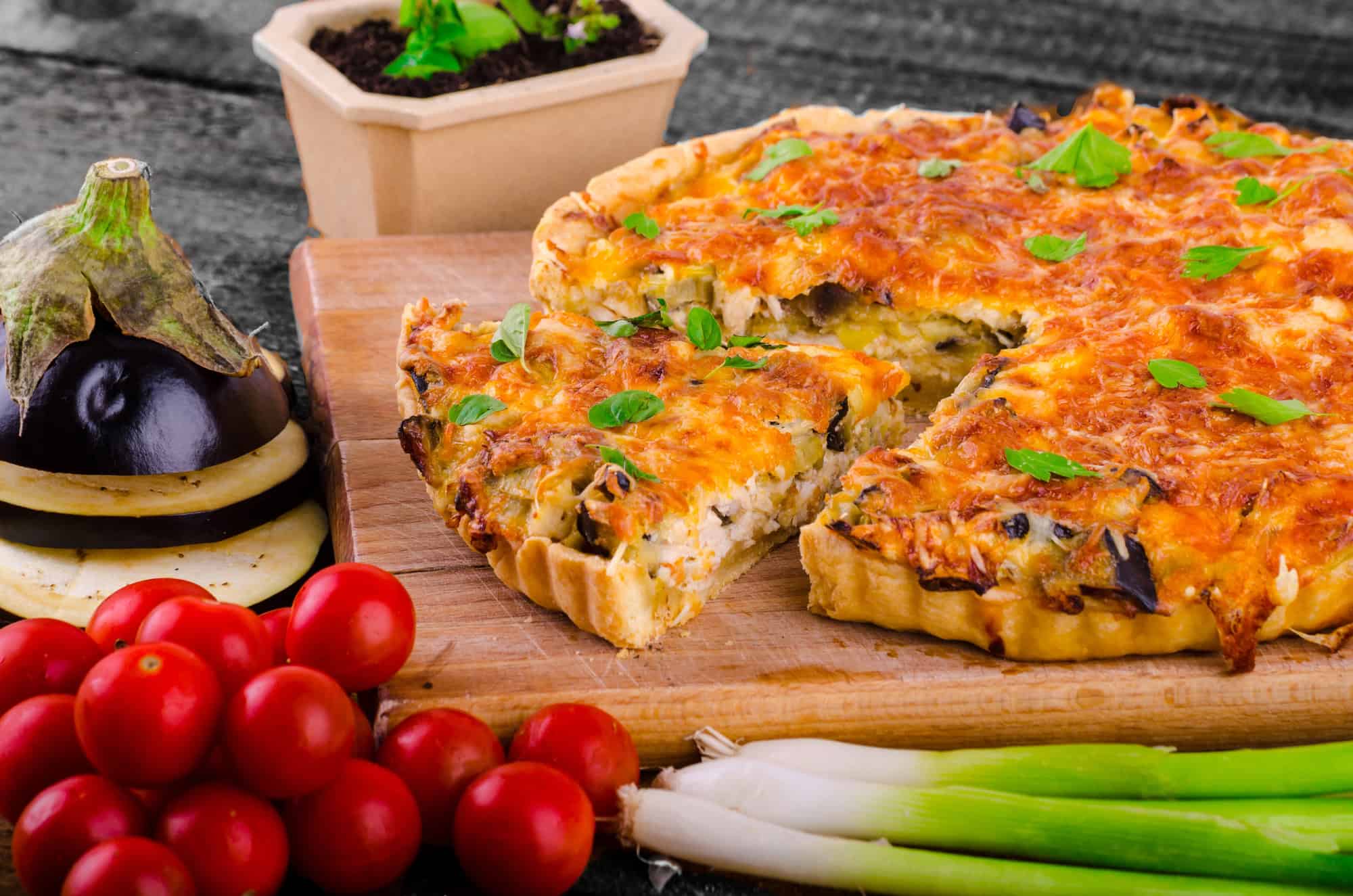 A vegetable quiche with herbs, surrounded by cherry tomatoes, eggplant, and green onions on a wooden board&mdash;a delicious example of Canadian Vegetarian Dishes.