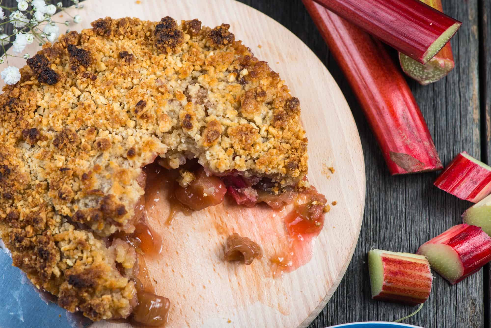 A crumb-topped rhubarb pie, inspired by Canadian Vegetarian Dishes, sits on a wooden board with a slice missing, next to fresh rhubarb stalks.