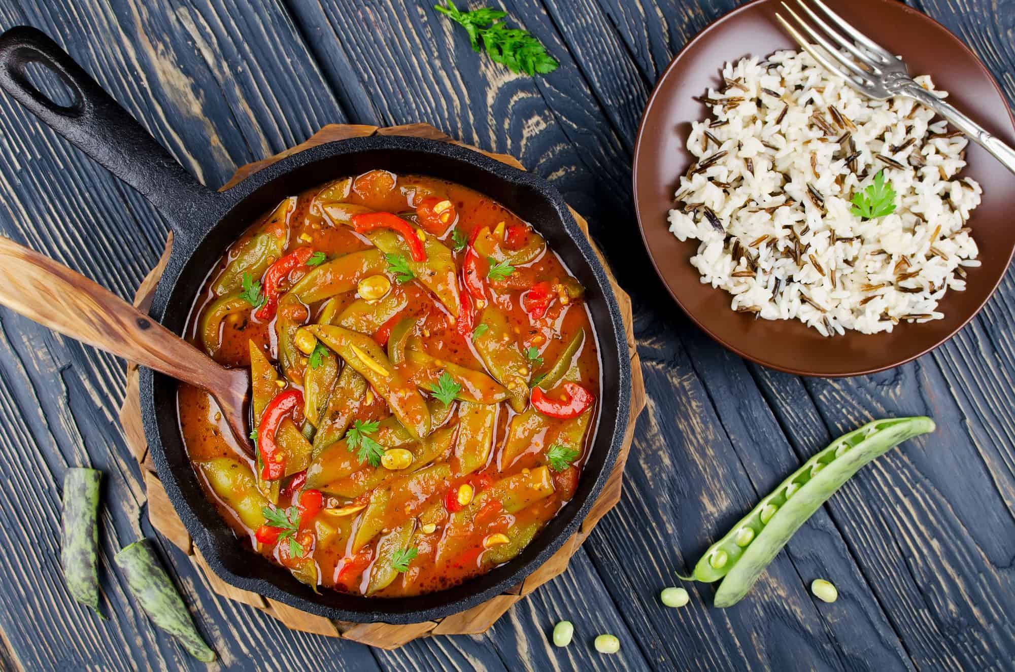 A skillet of vegetable curry, inspired by Canadian vegetarian dishes, with a wooden spoon next to a plate of cooked rice on a wooden table.